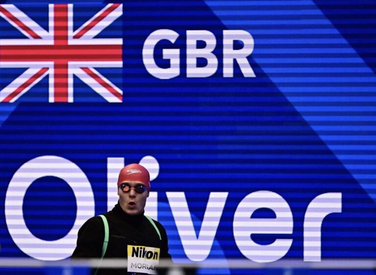 Oliver Morgan standing in front of a Great Britain sign wearing a swim cap and goggles.