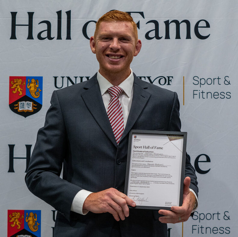 James Rodwell stood against a Hall of Fame background holding a certificate in his hand.