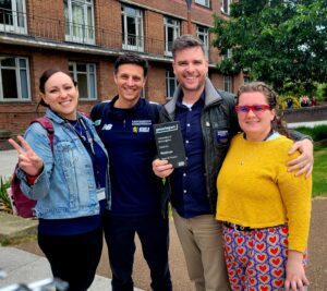 Four staff members stand in a line with arms round each other, holding the grey slate award, which reads 'Platinum Award'