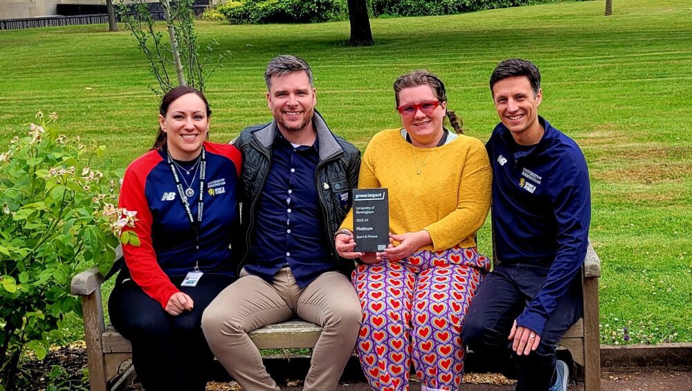 Four members of the UoB Sport & Fitness team sit on a bench in the Green Heart at UoB, holding the Platinum award