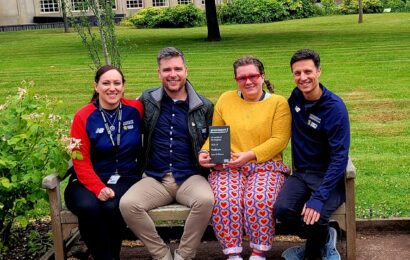 Four members of the UoB Sport & Fitness team sit on a bench in the Green Heart at UoB, holding the Platinum award